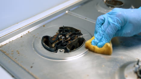 Scrubbing a greasy stove. A hand wearing a blue glove scrubs a dirty stovetop with a sponge, focusing on grease removal in the kitchen