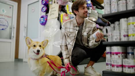 Confident-brunette-guy-in-a-light-checkered-shirt-chooses-food-for-his-yellow-corgi-dog-in-a-pet-store.-Confident-brunette-guy-with-his-dog-in-a-pet-store