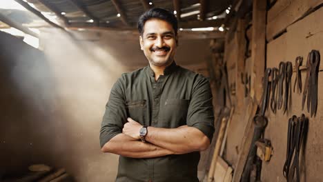 proud indian carpenter smiling with crossed arms in his messy workshop, dusty light is coming from the roof, many tools are hanging on the wall