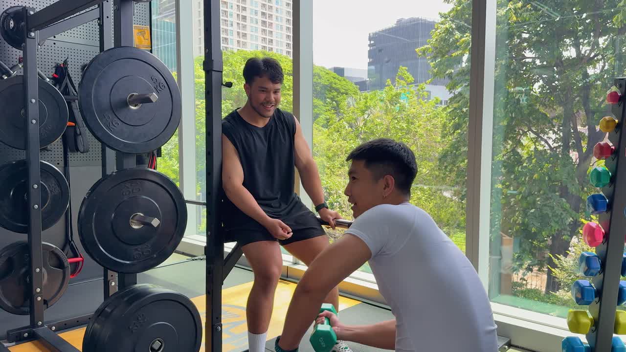 Two men relax and talk by weight racks in a bright gym with city views