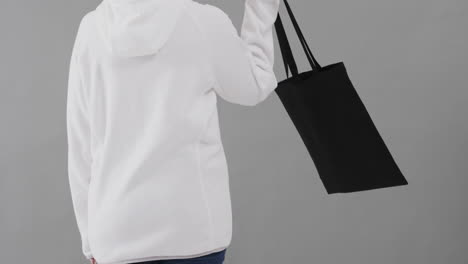 una mujer caucásica con una sudadera blanca con capucha sosteniendo una bolsa negra sobre un fondo gris, copia el espacio, cámara lenta.