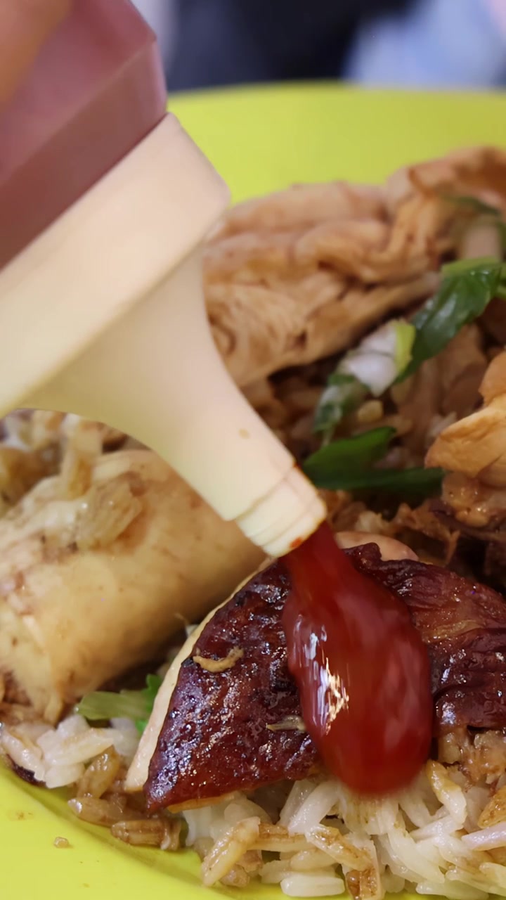 Una mano spreme salsa chili su riso con pollo arrosto, salsiccia e cipollotti su un piatto luminoso, sotto luce naturale con una prospettiva ravvicinata