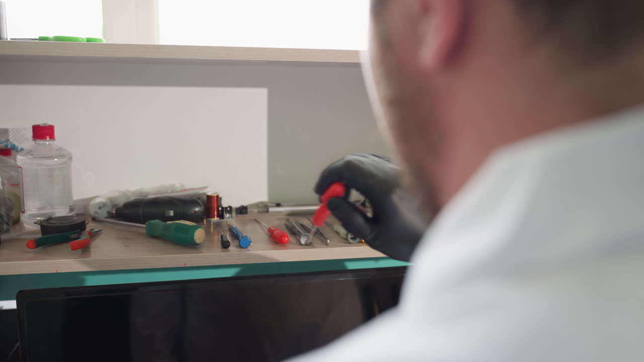 vista cercana de un hombre con una bata de laboratorio alcanzando un destornillador rojo en un estante, con varios equipos y herramientas de laboratorio en primer plano