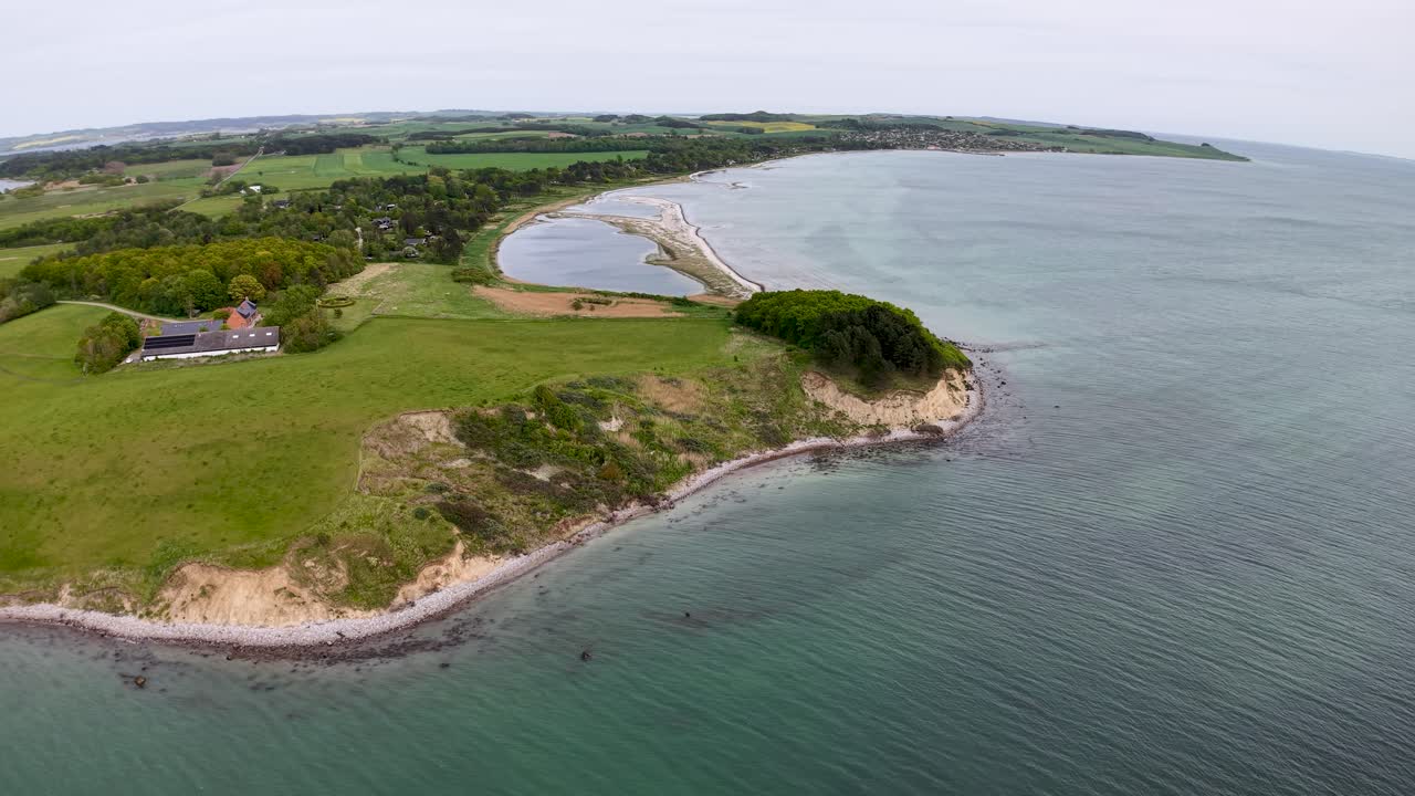 Aerial drone footage of a Danish peninsula surrounded by calm blue water, featuring green fields, trees, and a curved shoreline under soft daylight