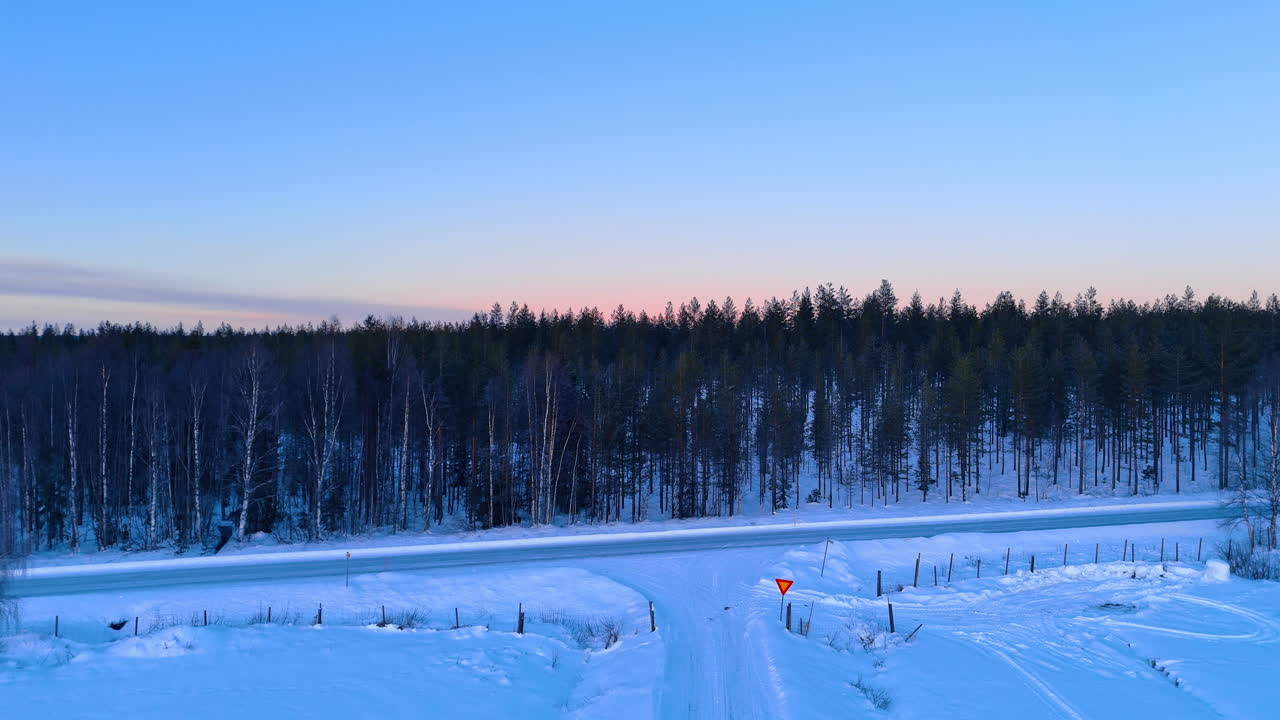Snødekt vei som fører til tett furuskog i Lapplands vinter. Vintervei i Lappland med snødekte jorder og furuskog under blå skumringshimmel med et veiskilt