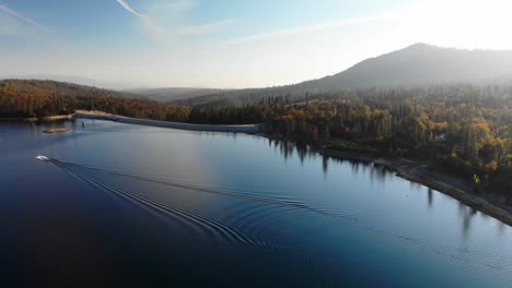 aerial shot of a dam on a blue alpine lake surrounded by pine trees, mountains, and cloudy sky