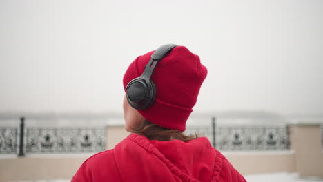 vista posteriore di una donna che indossa un berretto rosso e un cappuccio che ascolta le cuffie, guardando nel lontano orizzonte nebbioso durante l'inverno, lo sfondo presenta una recinzione decorativa e un ambiente nebbioso atmosferico