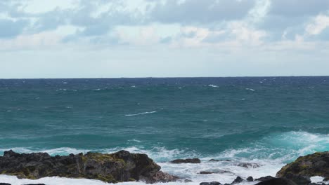 wide view of turquoise ocean waves approaching a rocky shoreline, white surf breaking against jagged black rocks under a bright yet cloudy sky