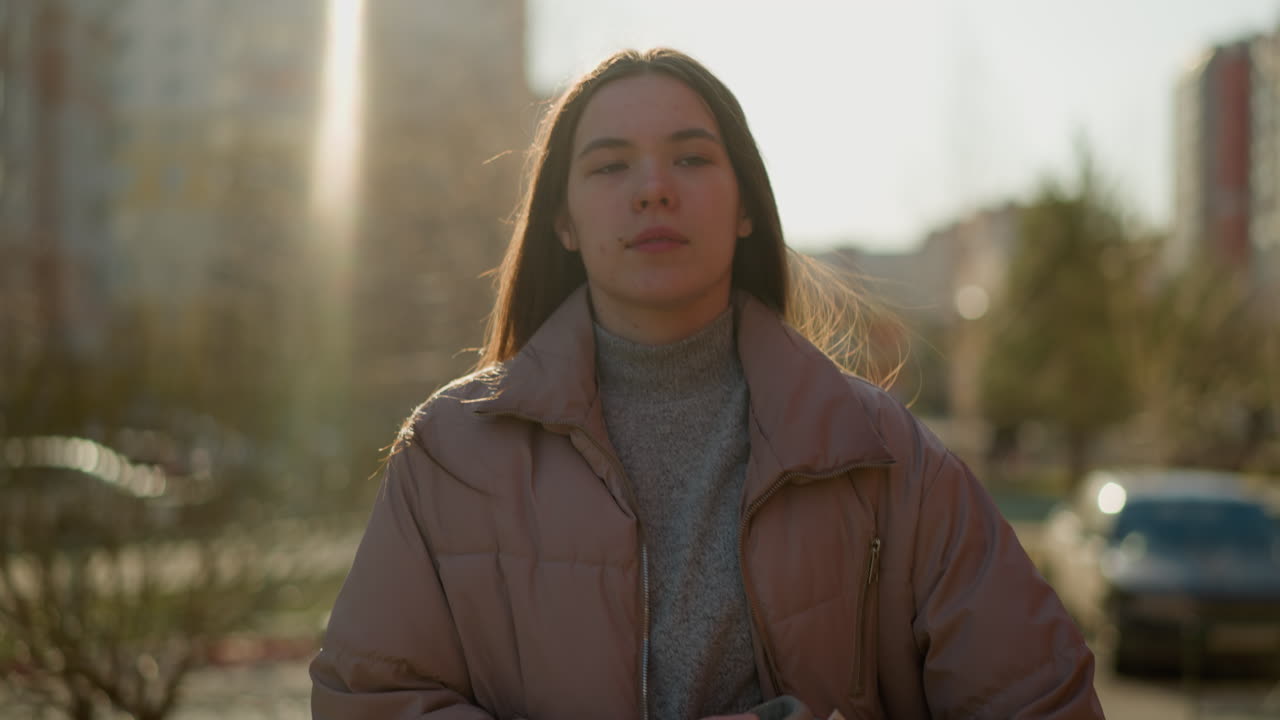 Front view of a girl walking through an urban area at sunset, wearing a peach jacket and a grey inner shirt. Her mouth is slightly open, and she gazes forward with a calm expression