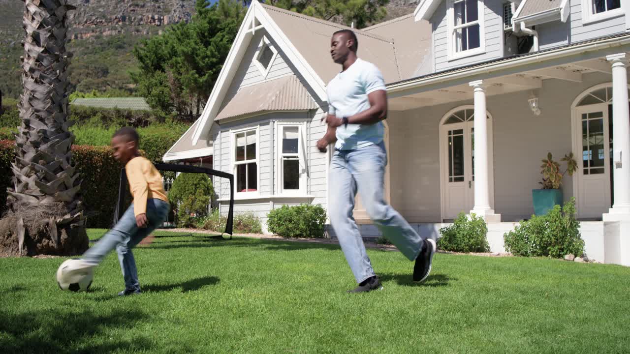 Pai e filho afro-americanos caminhando em direção à bola de futebol, chutando e defendendo perto de um gol portátil.