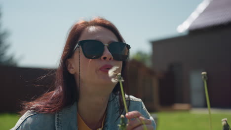 close up of delicate dandelion with white cotton seeds ready to disperse, with woman bending in background preparing to blow on it, set in sunny green backyard with house and garden in soft blur