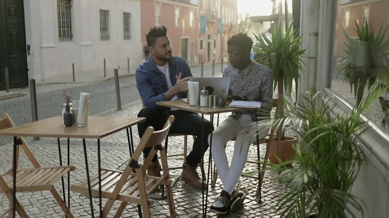 hombres multirraciales con dispositivos digitales hablando en un café al aire libre