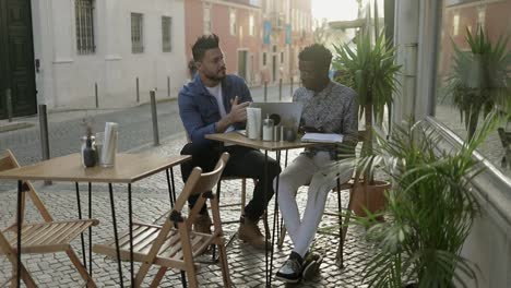 multiracial men with digital devices talking in outdoor cafe