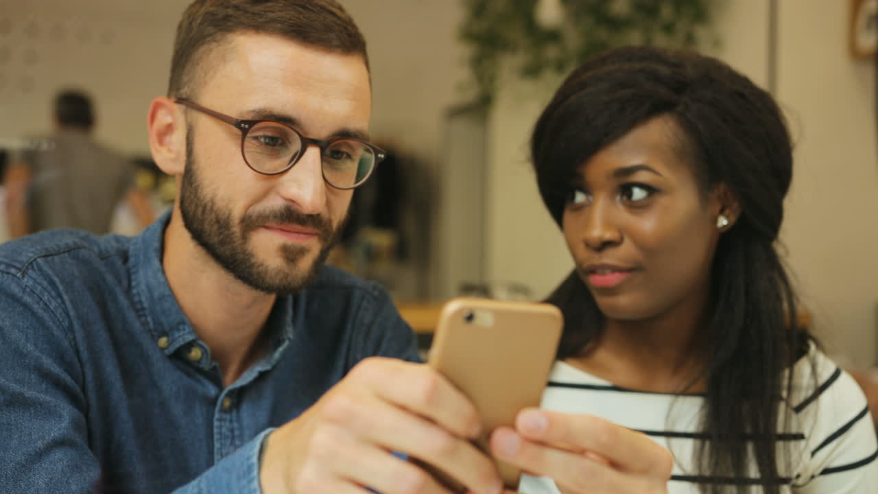 Close-up view of young caucasian man using smartphone and showing a video to african american woman a in a coffee shop