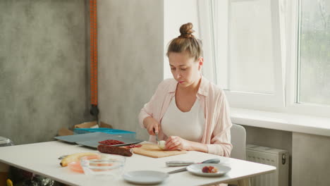 elegant young lady seated by bright window, carefully slicing onion on wooden board with sliced sausage, banana, boiled eggs and plates arranged on white table for home cooking preparation scene