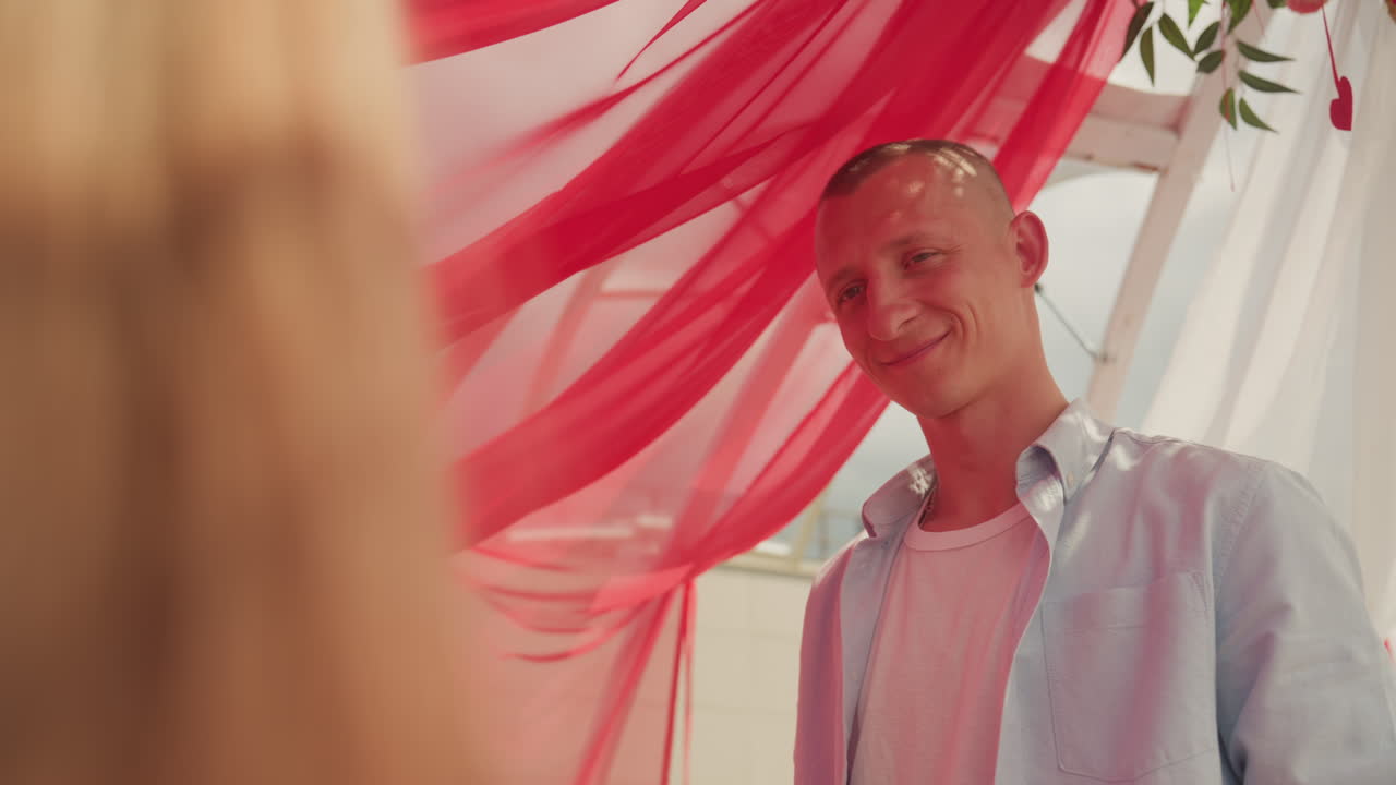 Homem sorrindo sob um toldo vermelho transparente, compartilhando um momento espontâneo e caloroso com um(a) parceiro(a) não visível, luz solar suave filtrando-se através das cortinas ondulantes, camisa casual e vibe relaxante de festival, decoração boêmia com tema floral