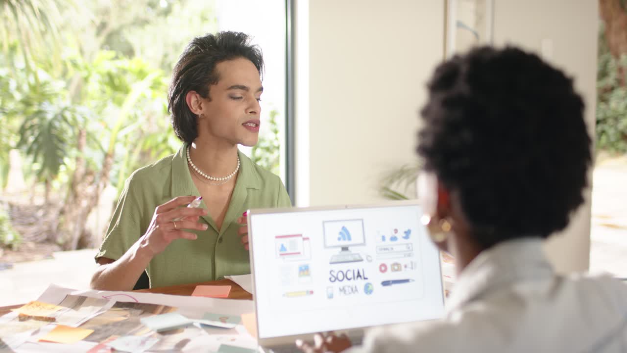 Diverse coworkers exchanging sticky notes at desk refining social graphics displaying on laptop