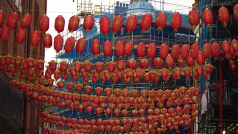 plenty of red lanterns hanged in china town in the middle of the street above windy weather moving flying around construction with blue material wrapped around repairing building in the background