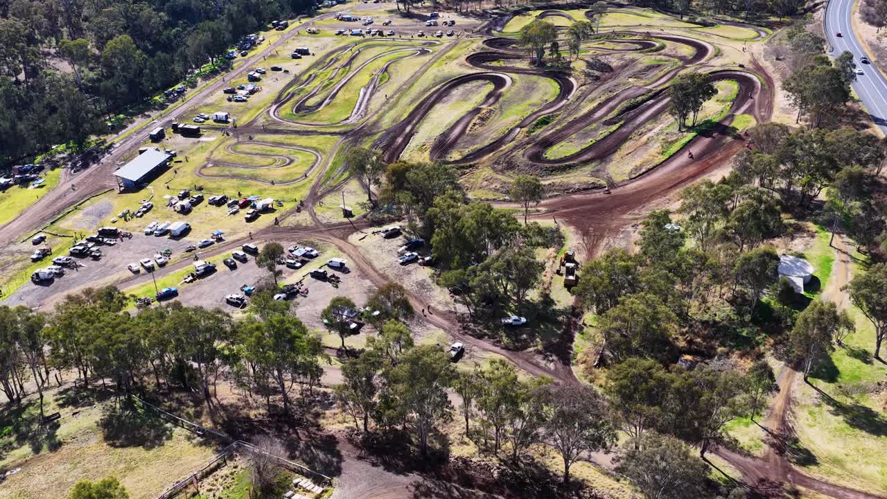 Drone footage captures a motocross event on a winding dirt track surrounded by eucalyptus trees, parked vehicles, and spectators under bright daylight