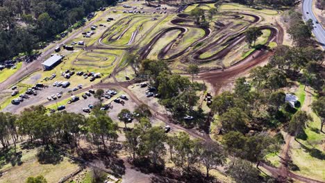 drone footage captures a motocross event on a winding dirt track surrounded by eucalyptus trees, parked vehicles, and spectators under bright daylight
