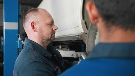 close-up head view of an engineer with earrings attentively watching a colleague using a pneumatic tool to tighten the bolt of a car in a garage