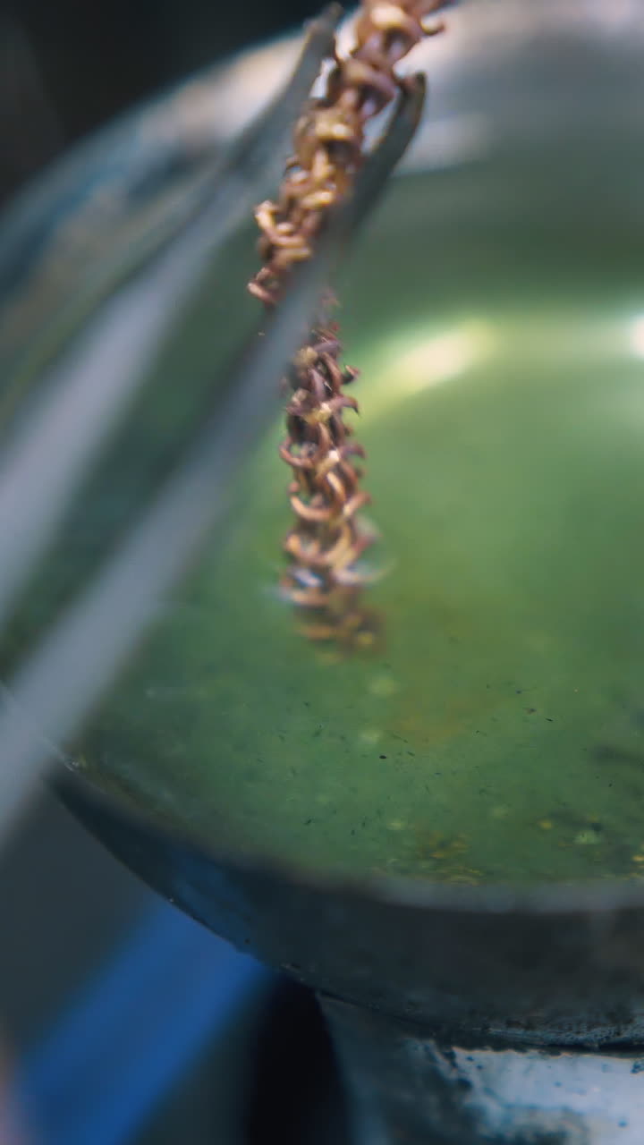 CU: professional jeweler puts gold chain into boiling green liquid in little bowl in light workshop extreme close view