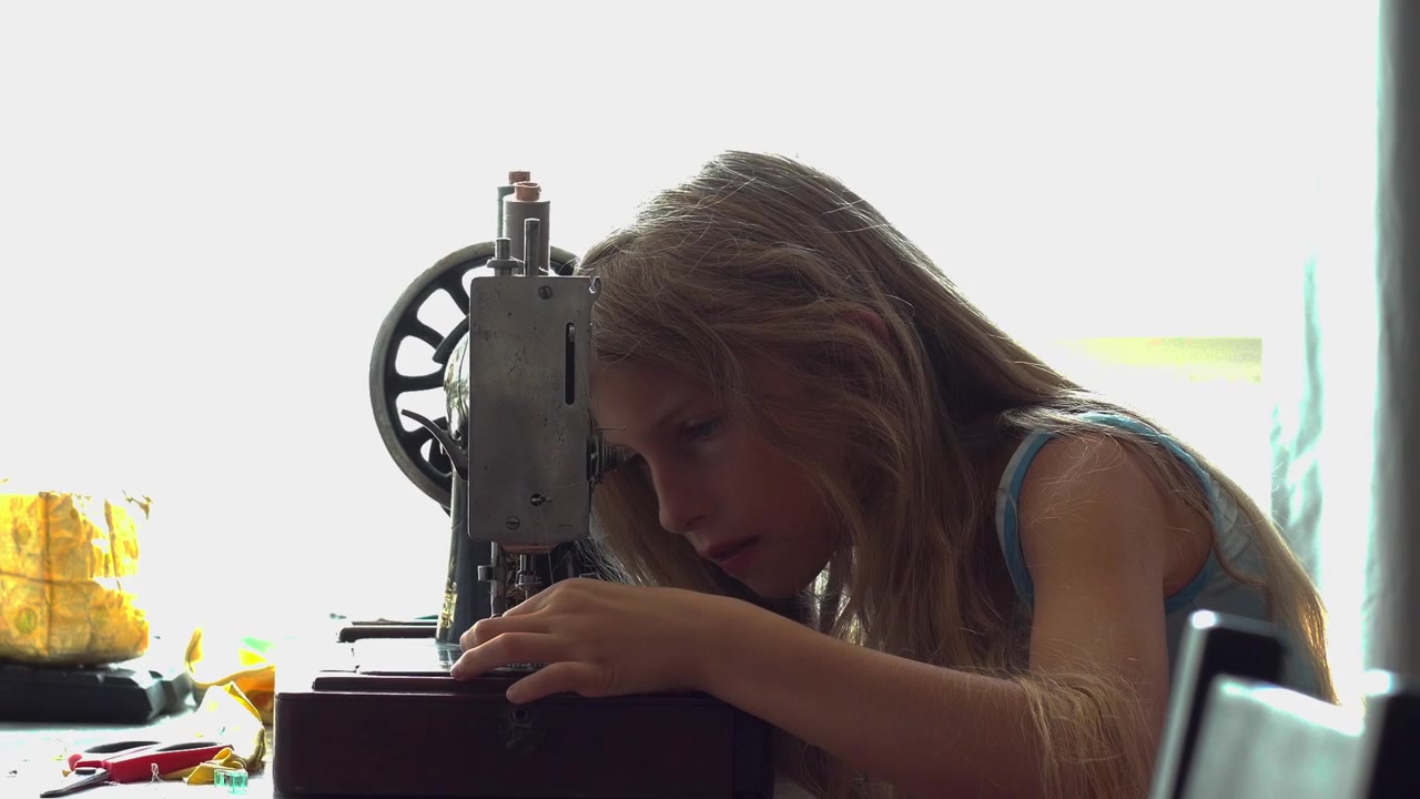 A medium static profile camera shot captures a young blonde girl sitting at a table carefully threading a needle on an antique mechanical sewing machine during a craft lesson in Budapest Hungary.