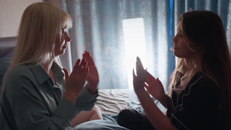 Side view of girls sitting on bed clapping hands together while playing traditional hand game indoors, expressing joy and connection as light from window