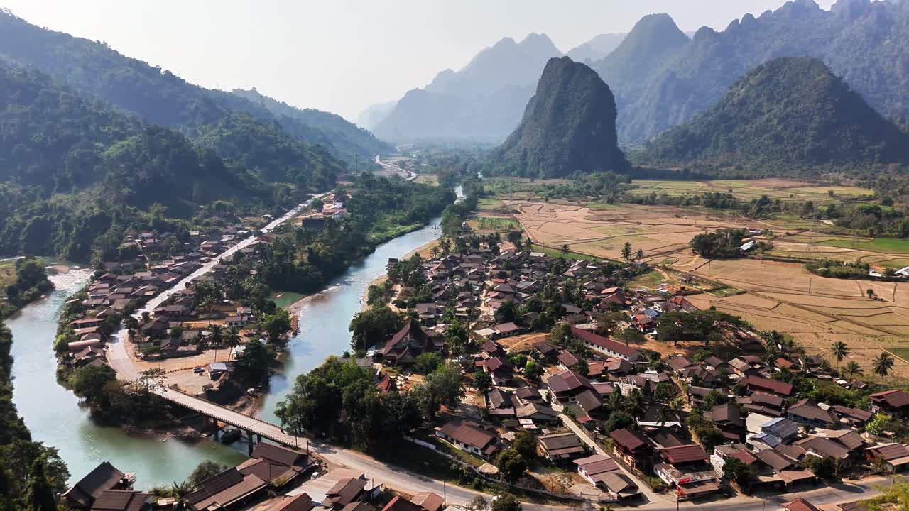 Aerial orbit of Pha Tang village nestled between limestone karst peaks, lush farmland, and the Nam Song River near Vang Vieng in Laos, showing the scenic rural valley from above