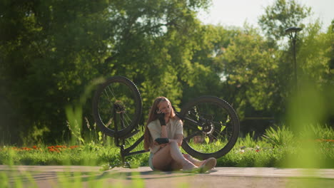 lady in black gloves seated next to her bike kept upside down, answering phone call, with vibrant greenery and trees in the background