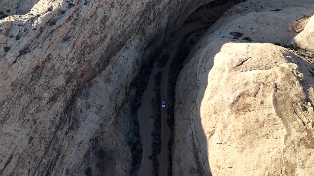 Luftfoto av hvit SUV som kjører i en ørkenkløft under bratte sandsteinsklipper, avslørende droneopptak