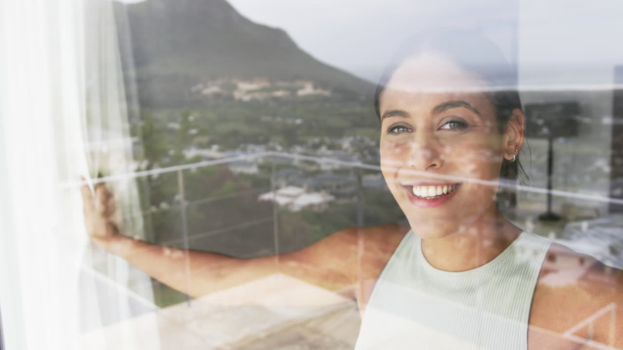 retrato de una feliz mujer biracial mirando por la ventana de su casa, en cámara lenta