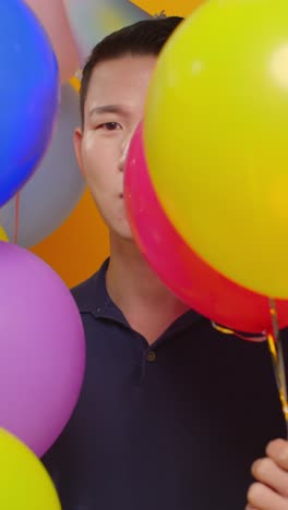 파티 모자를 입은 남자가 풍선 뒤에 숨어 생일을 축하하는 수직 비디오 스튜디오 초상화