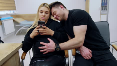 pregnant blonde woman sits looking at her smartphone and talking. her leans on her shoulder and pets her belly.