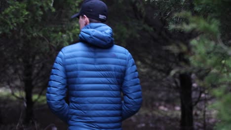 Young-white-male-in-a-blue-jacket-and-black-ball-cap-walking-in-a-forest-in-Kentucky-and-looking-around-curious