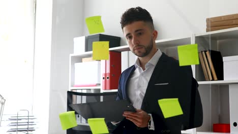 hombre de negocios en abrigo de traje haciendo notas en documentos de pie cerca de la pared de vidrio