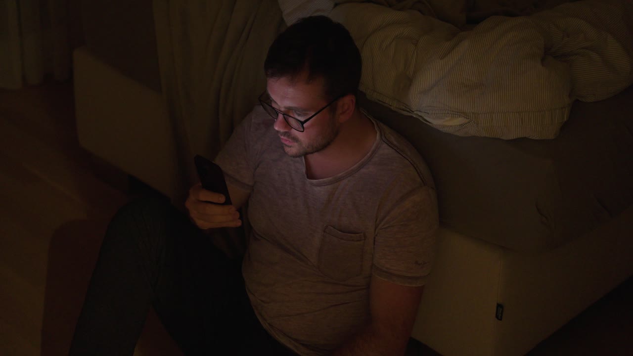 Un uomo seduto sul pavimento in una stanza buia, illuminato dallo schermo del suo telefono, simboleggia la dipendenza dai social media e le cattive abitudini notturne legate all'uso dello schermo.