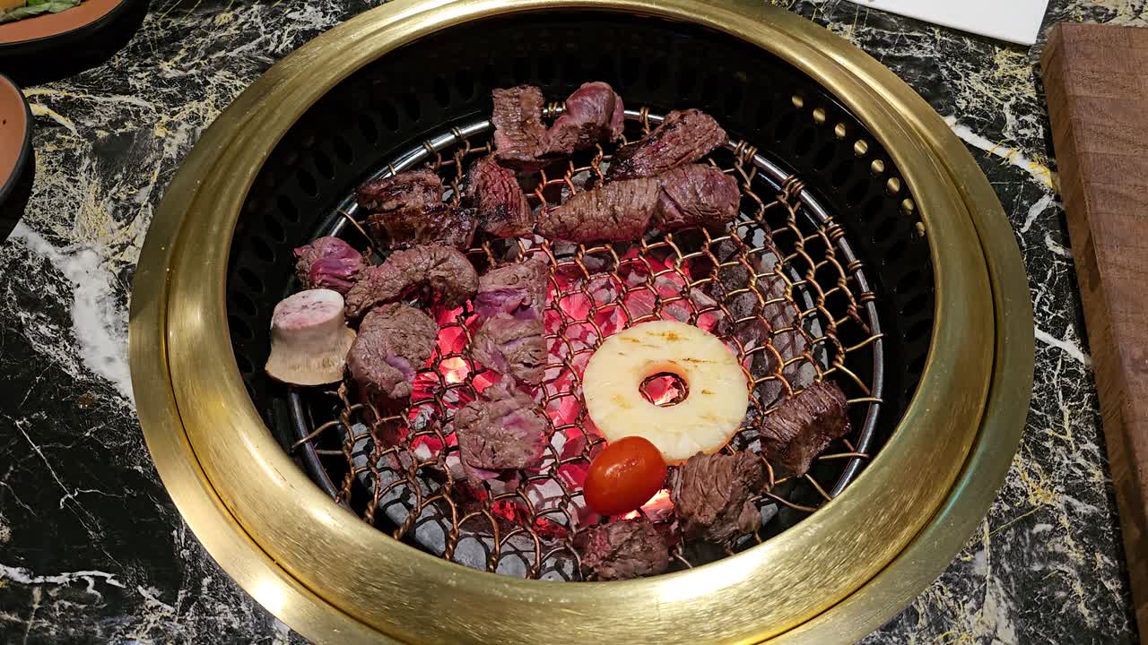 A chef expertly uses tongs to flip sizzling beef, pineapple, and vegetables on a glowing grill at Hanu Beef Korean restaurant, showcasing a lively Korean barbecue experience - top down view