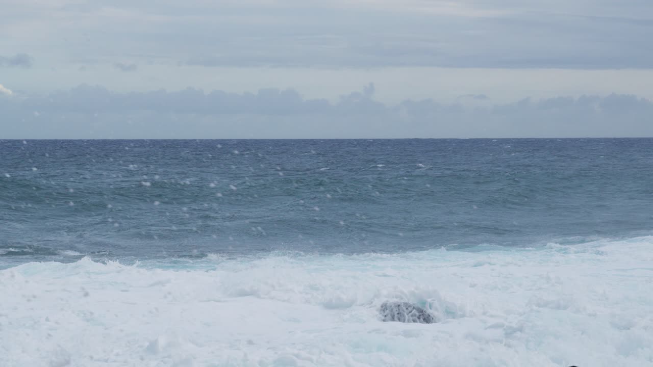 Foamy white surf lifts and falls along a rocky shoreline while a steel-blue horizon breathes under gray clouds, a steady, meditative seascape of motion, weather, and untamed energy