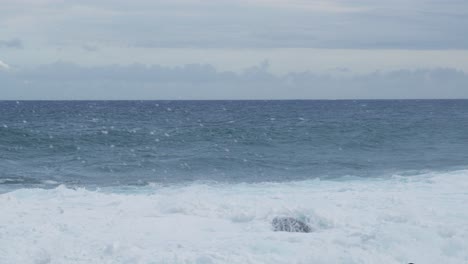 foamy white surf lifts and falls along a rocky shoreline while a steel-blue horizon breathes under gray clouds, a steady, meditative seascape of motion, weather, and untamed energy