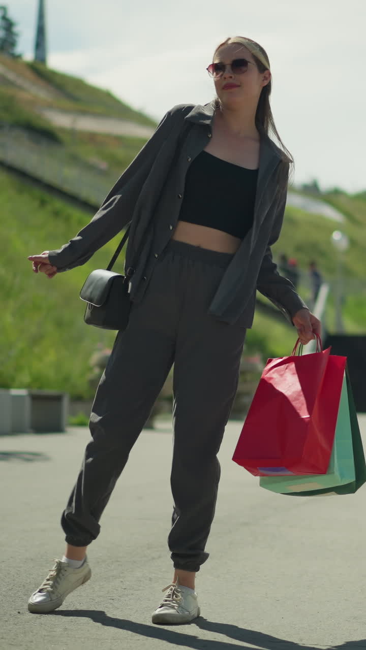 dama con gafas de sol baila felizmente con bolsas de compras en su mano derecha, su sombra es visible en el suelo, con una colina a un lado, la gente camina cerca de una escalera en el fondo