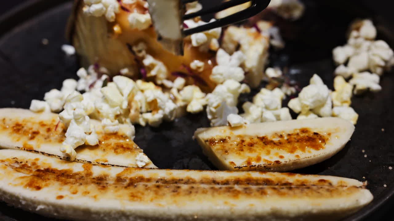 Close up of a woman cutting a cheesecake with a banana split and caramelised popcorn on a black plate at a restaurant