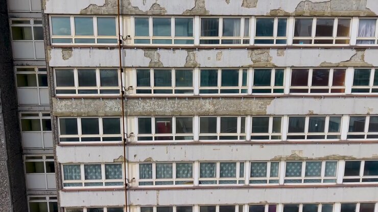 Un condominio fatiscente a Stratford si innalza con un movimento ascendente del drone, rivelando vernice scrostata, intonaco crepato, fasce di cemento macchiate e file di finestre invecchiate su un esterno trascurato di un grattacielo