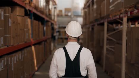 Rear-view-of-a-man-in-work-uniform-with-a-flashlight-walking-down-a-warehouse-corridor-with-boxes