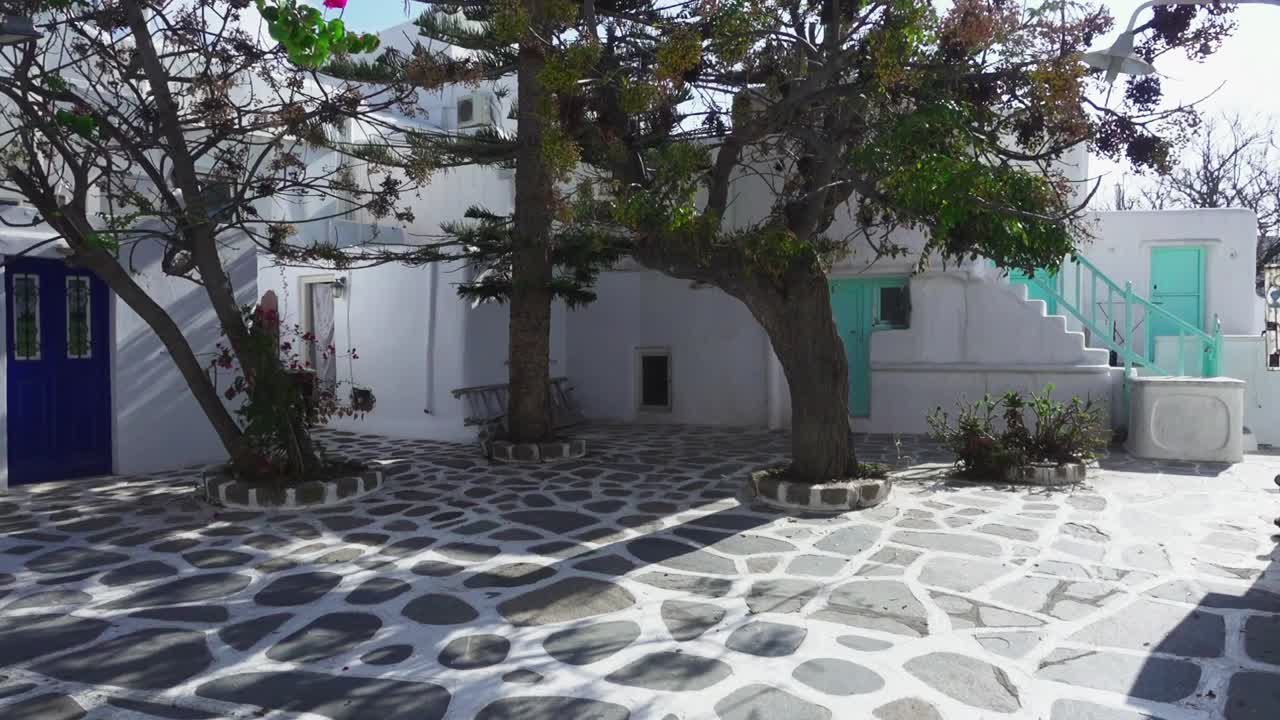 Sliding view of an empty, tiled courtyard with trees casting shadows in front of typical white, Greek houses on the island of Mykonos