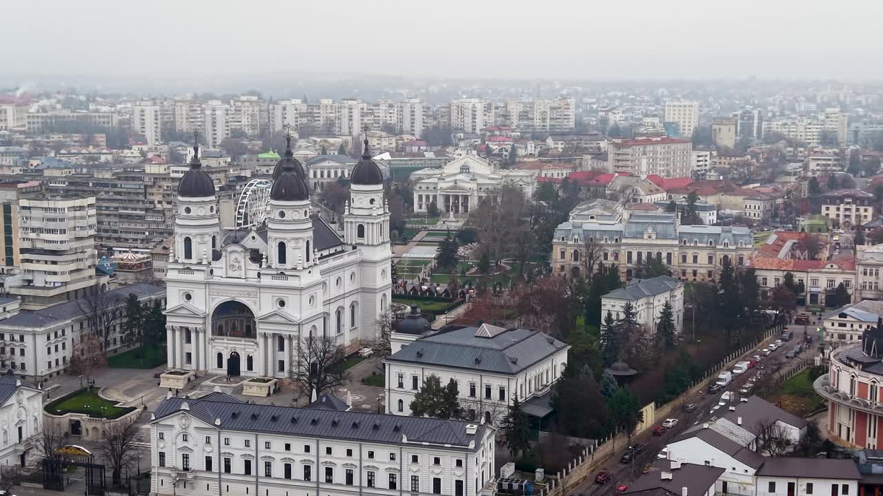 Vídeo de drone da Catedral Metropolitana no centro da cidade de Iasi, Romênia, mostrando um edifício branco com quatro cúpulas e torres pretas cercado por quarteirões urbanos e área de parque verde em um dia nublado