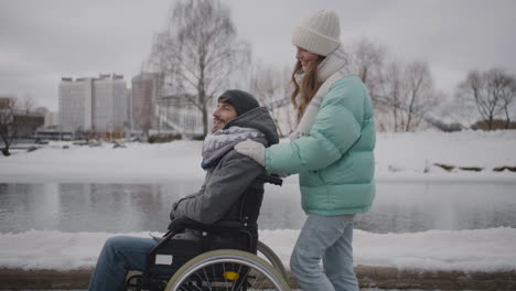 side view of happy caucasian woman taking her disabled friend in wheelchair for a walk in the winter city and talking together