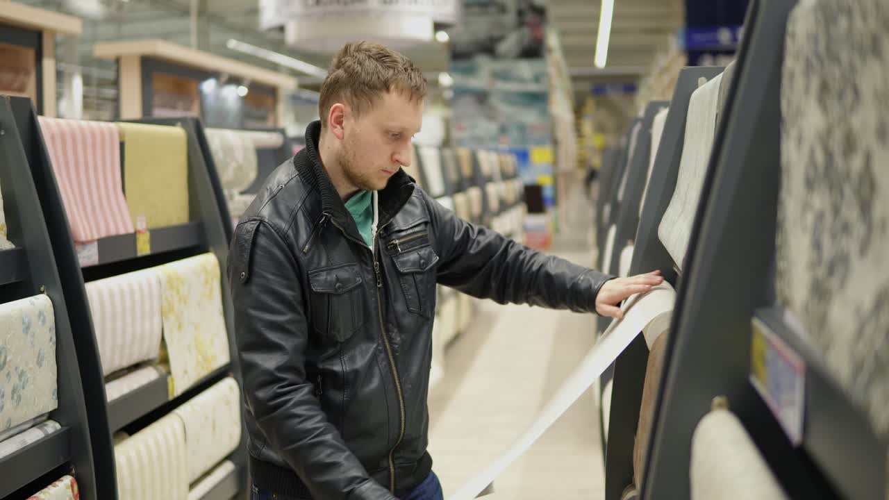 Young man choosing wallpapers in supermarket. He is looking at the wallpaper, touching it and thinking which one is better