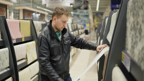 Junger-Mann-Wählt-Tapeten-Im-Supermarkt.-Er-Betrachtet-Die-Tapete,-Berührt-Sie-Und-überlegt,-Welche-Besser-Ist