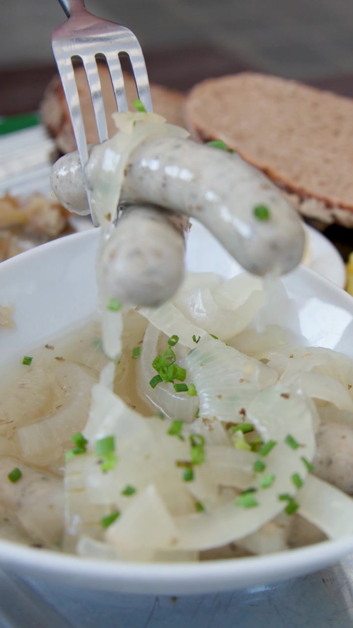 Una mano solleva una salsiccia di Norimberga da una ciotola di crauti e insalata di patate, con illuminazione brillante e naturale.